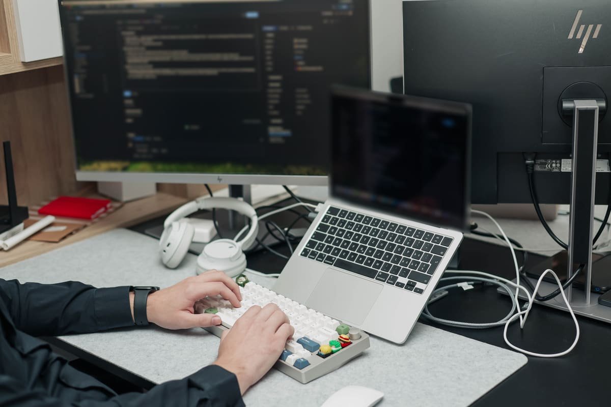 Hands on a mechanical keyboard in front of a laptop and monitor with code